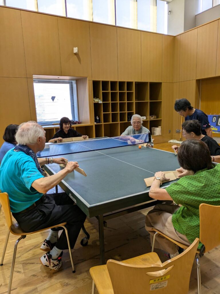 卓球台を囲み、いすに座って木のラケットで卓球バレーを楽しむ様子